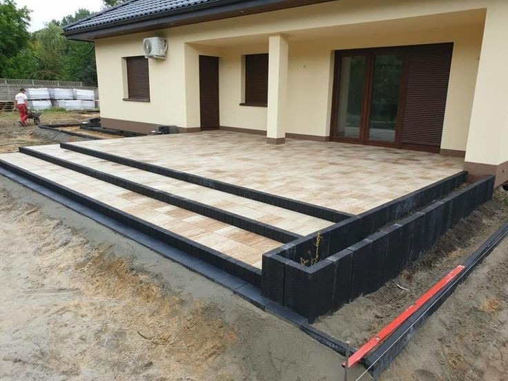 Wykończony taras z kostki brukowej, prowadzący do domu, z trzema stopniami i obrzeżami z ciemnych bloczków. W tle pracownik z taczką i palety z materiałem.