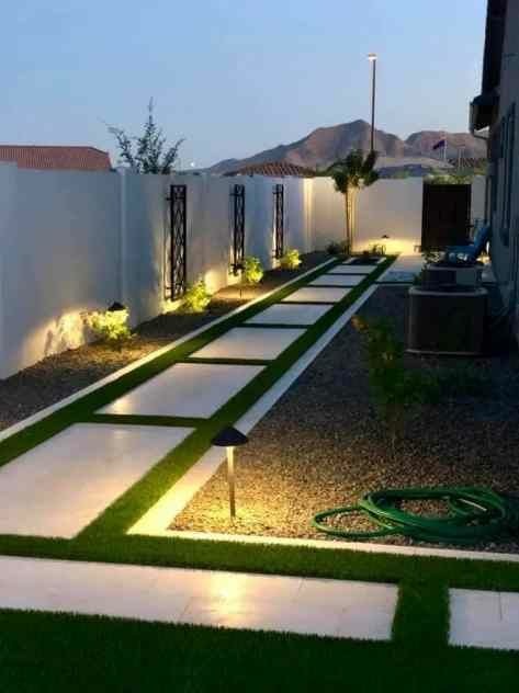 Oświetlona ścieżka z płyt betonowych rozdzielonych sztuczną trawą, prowadząca przez żwirowe podwórko, z dekoracyjnymi elementami na białym murze i lampkami w tle.