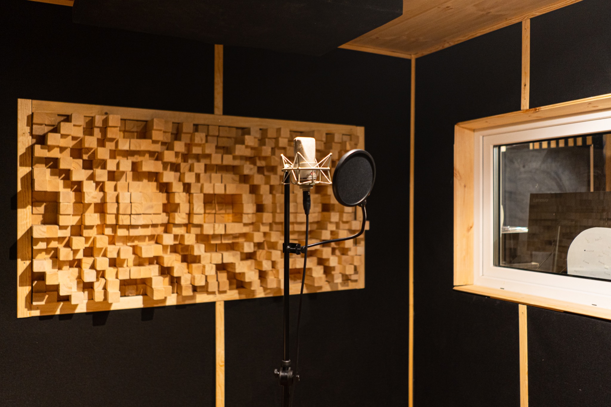 Profesjonalny mikrofon studyjny z pop filtrem na statywie, ustawiony przed drewnianym panelem akustycznym w pomieszczeniu wyizolowanym akustycznie z oknem.