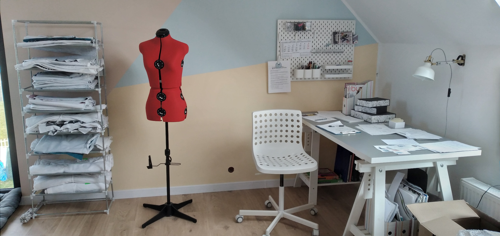 Pracownia krawiecka na poddaszu: czerwony manekin krawiecki na czarnym statywie, regał z ułożonymi materiałami, biurko z dokumentami i białe krzesło na kółkach.