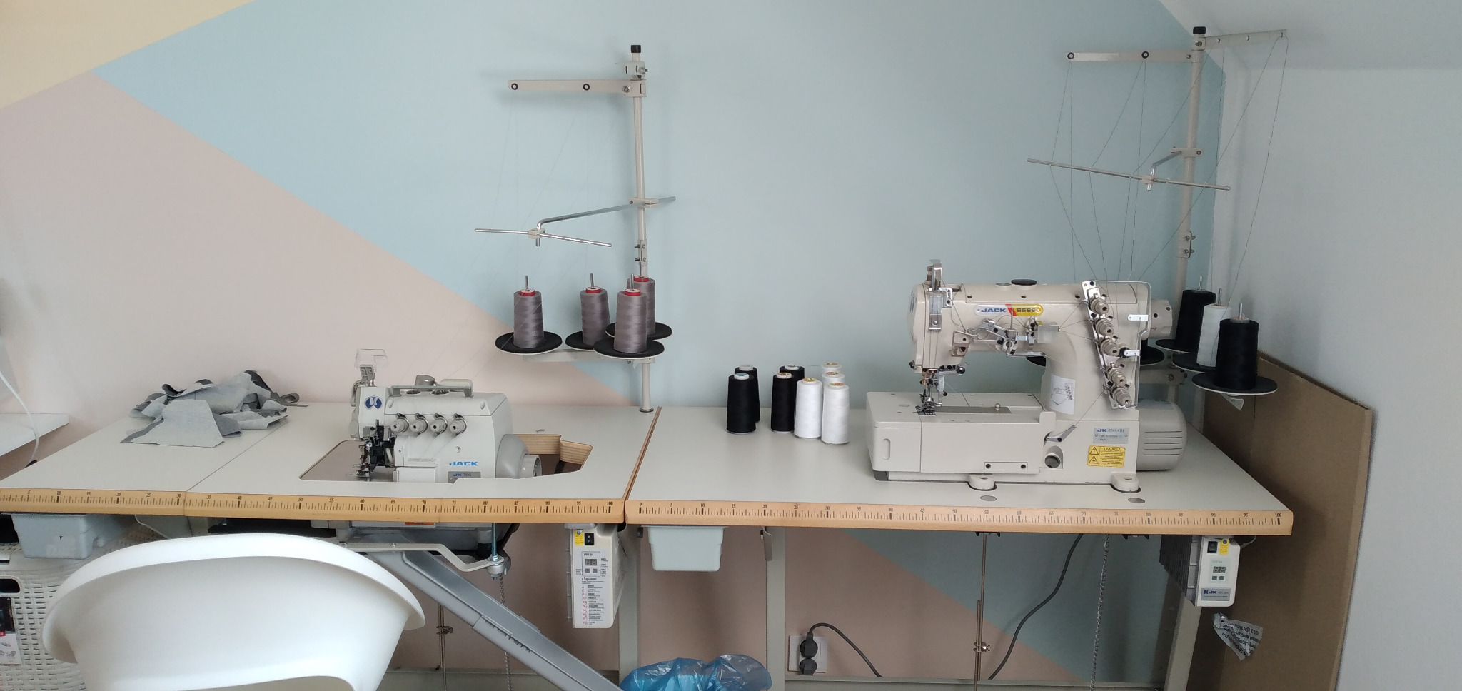 Dwie maszyny do szycia na białych stołach w pomieszczeniu z geometrycznie pomalowaną ścianą, widoczne szpule nici w różnych kolorach i fragmenty materiału.
