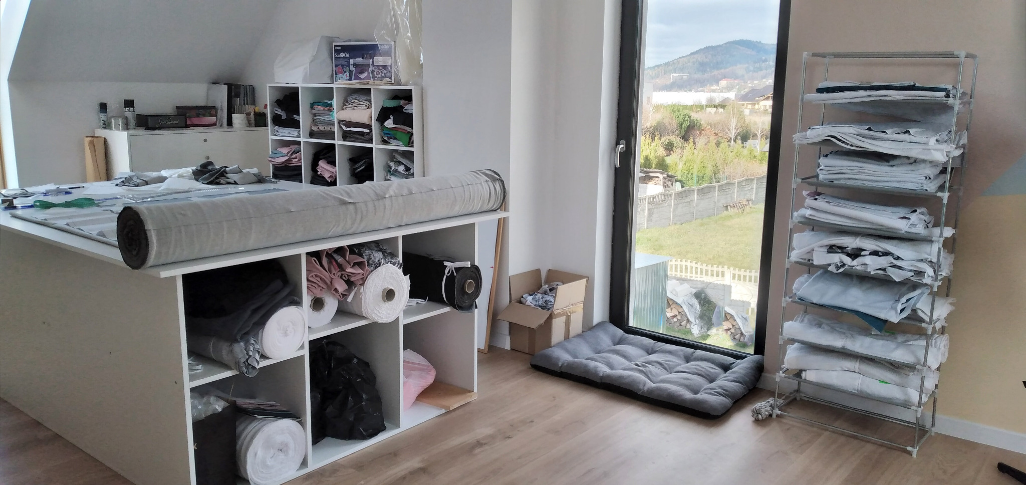 Pracownia krawiecka: rolka szarego materiału na stole krojczym, półki z tkaninami, regał z wykrojami, okno z widokiem na góry.