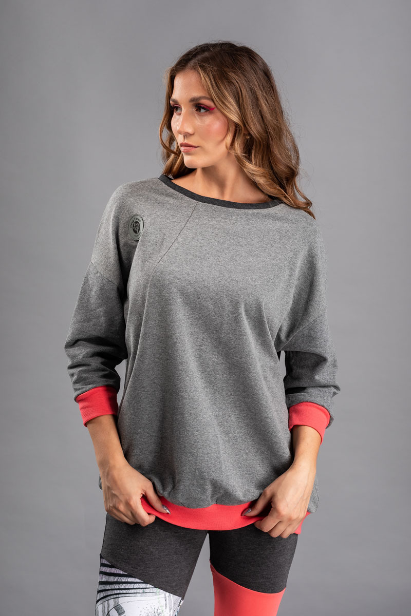 Modelka prezentuje szarą bluzę dresową z długimi rękawami i koralowymi mankietami oraz wykończeniem dołu, na jednolitym szarym tle.