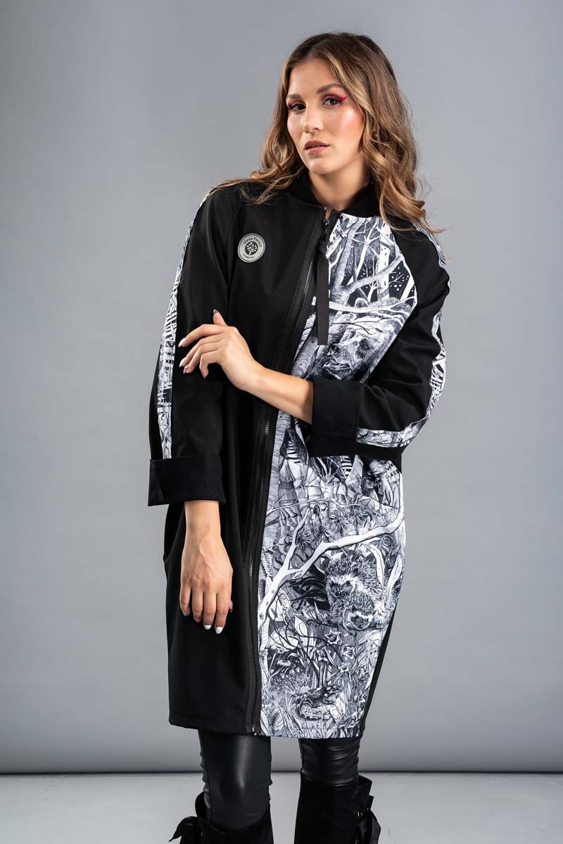Modelka prezentuje długi, czarno-biały płaszcz z asymetrycznym wzorem leśnym, z widocznym jeżem i drzewami, na szarym tle.