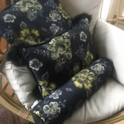 Komplet dekoracyjnych poduszek z ciemnego materiału w kwiatowy wzór, wykończonych czarnym futerkiem, ułożonych w wiklinowym fotelu z jasną poduchą.