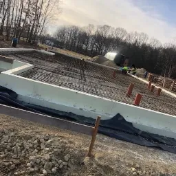 Przygotowanie zbrojenia fundamentów pod budowę domu jednorodzinnego, widoczne szalunki styropianowe, siatka zbrojeniowa, rury kanalizacyjne oraz pracownicy w oddali, tło stanowią drzewa i niebo.