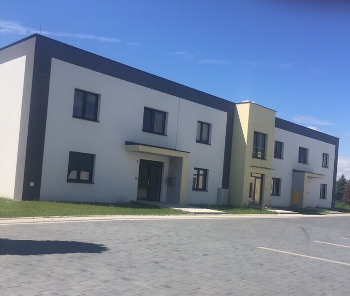 Nowoczesny budynek mieszkalny z płaskim dachem w Pszczynie, z szarą elewacją i żółtym akcentem przy wejściu, widok z poziomu parkingu z kostki brukowej.