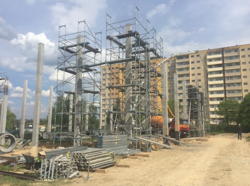 Wznoszenie konstrukcji szkieletowej budynku z prefabrykatów betonowych, widoczne rusztowania i betoniarka na tle bloku mieszkalnego.