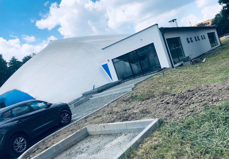 Nowoczesny budynek z białą elewacją i dużymi oknami, połączony z namiotową konstrukcją, widoczny samochód osobowy i teren wokół w trakcie prac wykończeniowych.