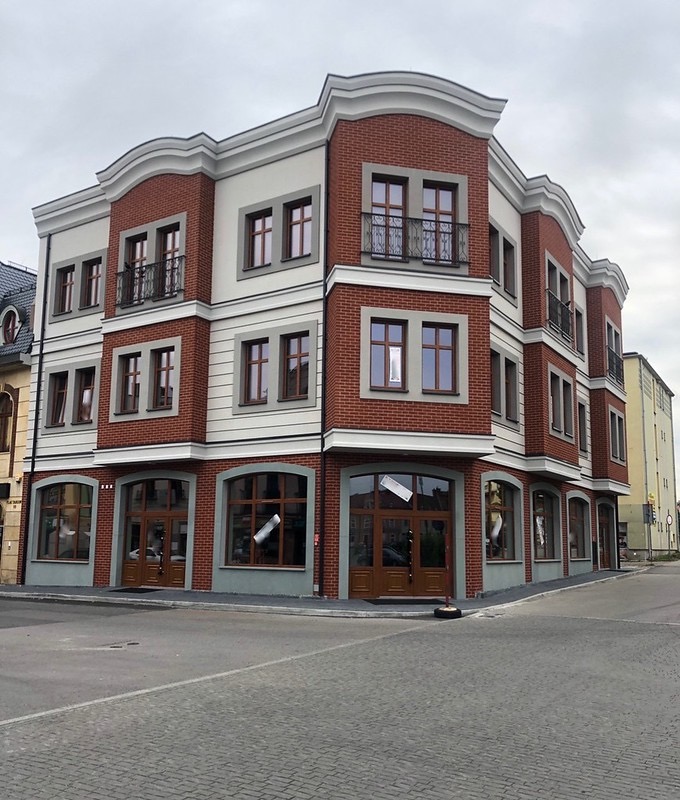 Nowo wybudowany, trzypiętrowy budynek narożny z czerwonej cegły z białymi elementami dekoracyjnymi, balkonami z kutymi balustradami i dużymi oknami na parterze, widoczny z poziomu ulicy wyłożonej...