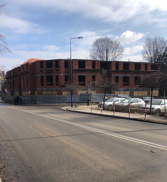 Wznoszenie dwukondygnacyjnego budynku z czerwonej cegły na rogu ulicy, zabezpieczonego szarą blachą falistą, widok z poziomu jezdni w pochmurny dzień.