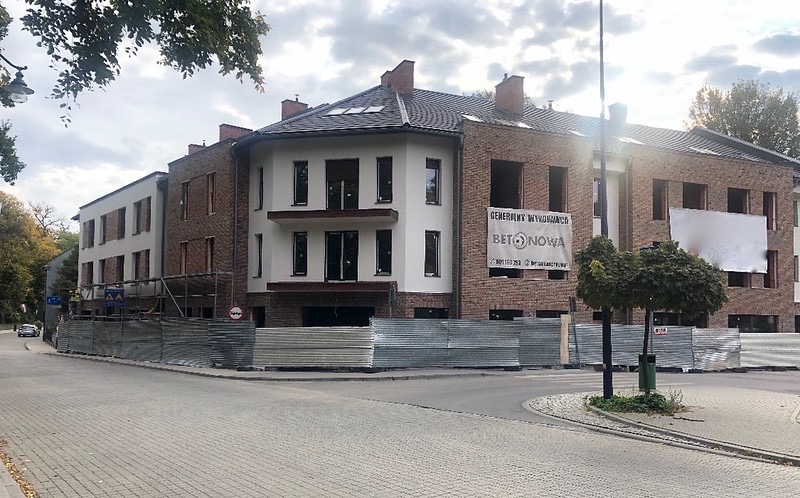 Budynek w trakcie budowy z ceglaną elewacją, widoczne rusztowania i ogrodzenie placu budowy, na budynku baner reklamowy firmy budowlanej.