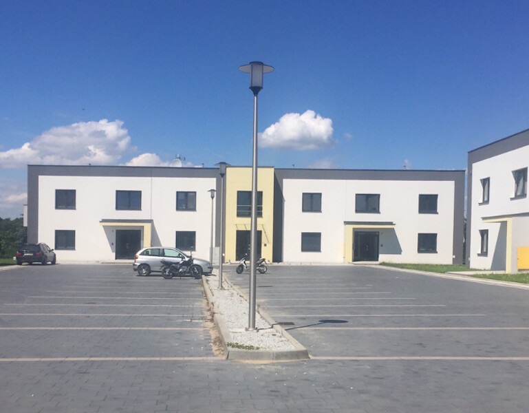 Nowoczesny budynek biurowy z parkingiem wyłożonym kostką brukową, biała elewacja z szarymi akcentami, błękitne niebo z chmurami w tle.