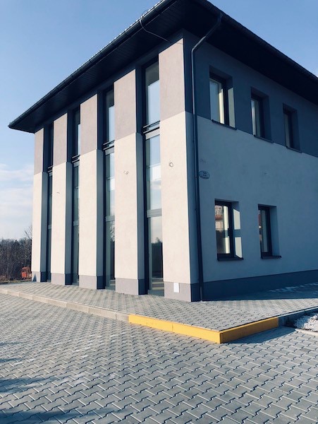 Dwukondygnacyjny budynek z szaro-białą elewacją i dużymi oknami, widoczny od strony chodnika wyłożonego szarą kostką brukową z żółtym obrzeżem.