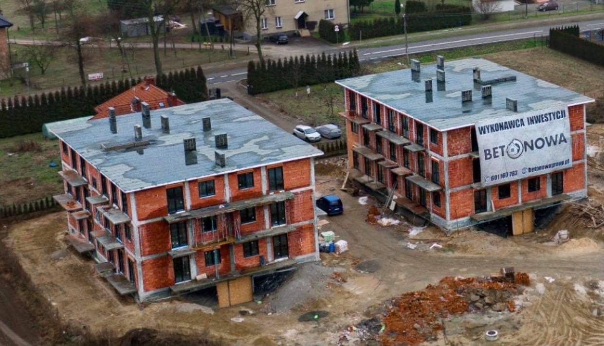 Dwa budynki wielorodzinne w stanie surowym otwartym, z widoczną konstrukcją z cegły i betonowymi stropami, na jednym z nich baner reklamowy firmy budowlanej.