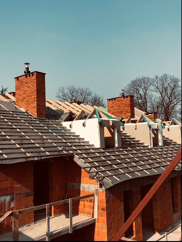 Budowa domu murowanego: widok na konstrukcję dachu z kominami w trakcie budowy, czerwona cegła, czarna membrana dachowa, drewniane elementy konstrukcyjne, niebieskie niebo w tle.