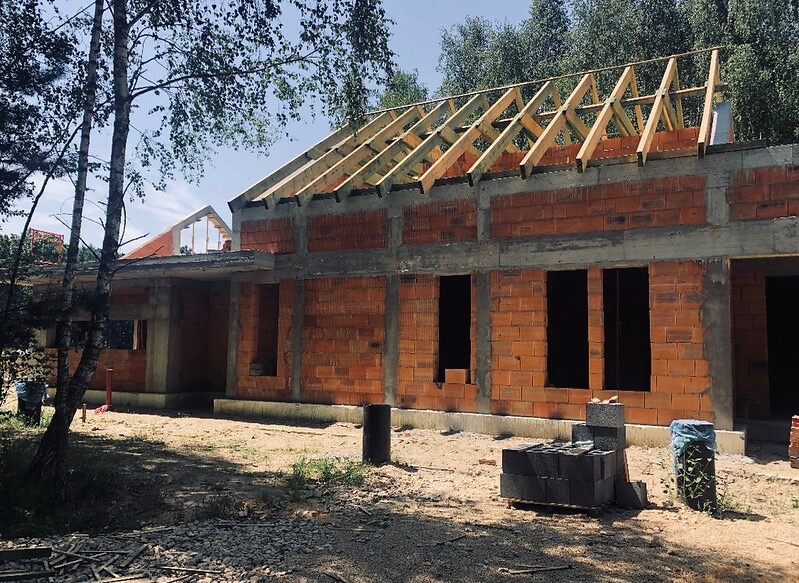 Budowa domu jednorodzinnego z czerwonej cegły, widoczne ściany i drewniana konstrukcja dachu na tle drzew.