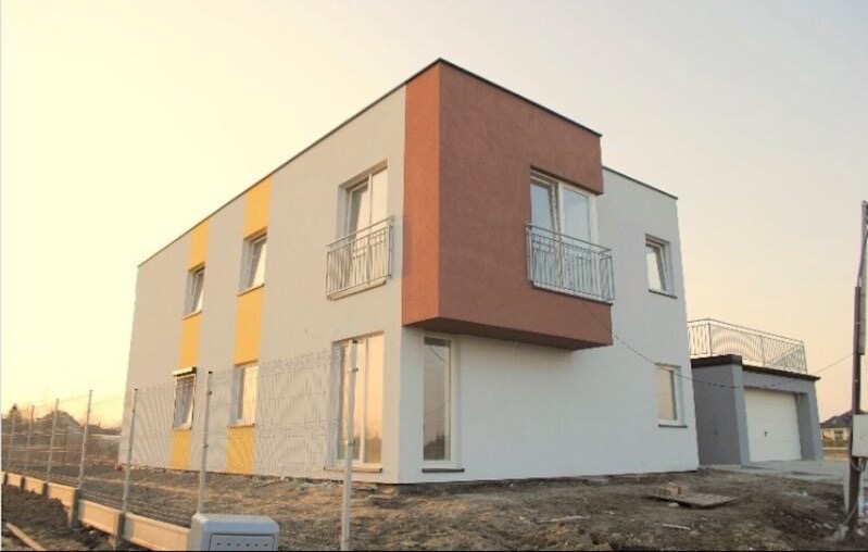 Nowoczesny, dwukondygnacyjny dom z płaskim dachem, wykończony tynkiem w kolorach białym, brązowym i żółtym, z balkonami z metalowymi balustradami i garażem w tle, ogrodzony siatką.