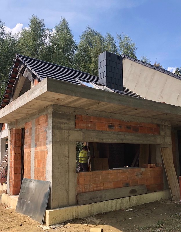 Surowy stan domu jednorodzinnego w trakcie budowy: widoczne ściany z czerwonej cegły, betonowe nadproża, fragment dachu z czarną dachówką i komin. W tle drzewa i pracownik w żółtej kamizelce.