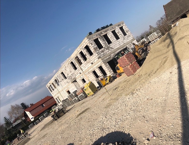 Wznoszenie dwukondygnacyjnego budynku z białych bloczków, widoczny żółty mini-ładowarka z paletą czerwonych cegieł, teren budowy z gruzem i piaskiem, w tle domy jednorodzinne i błękitne niebo.