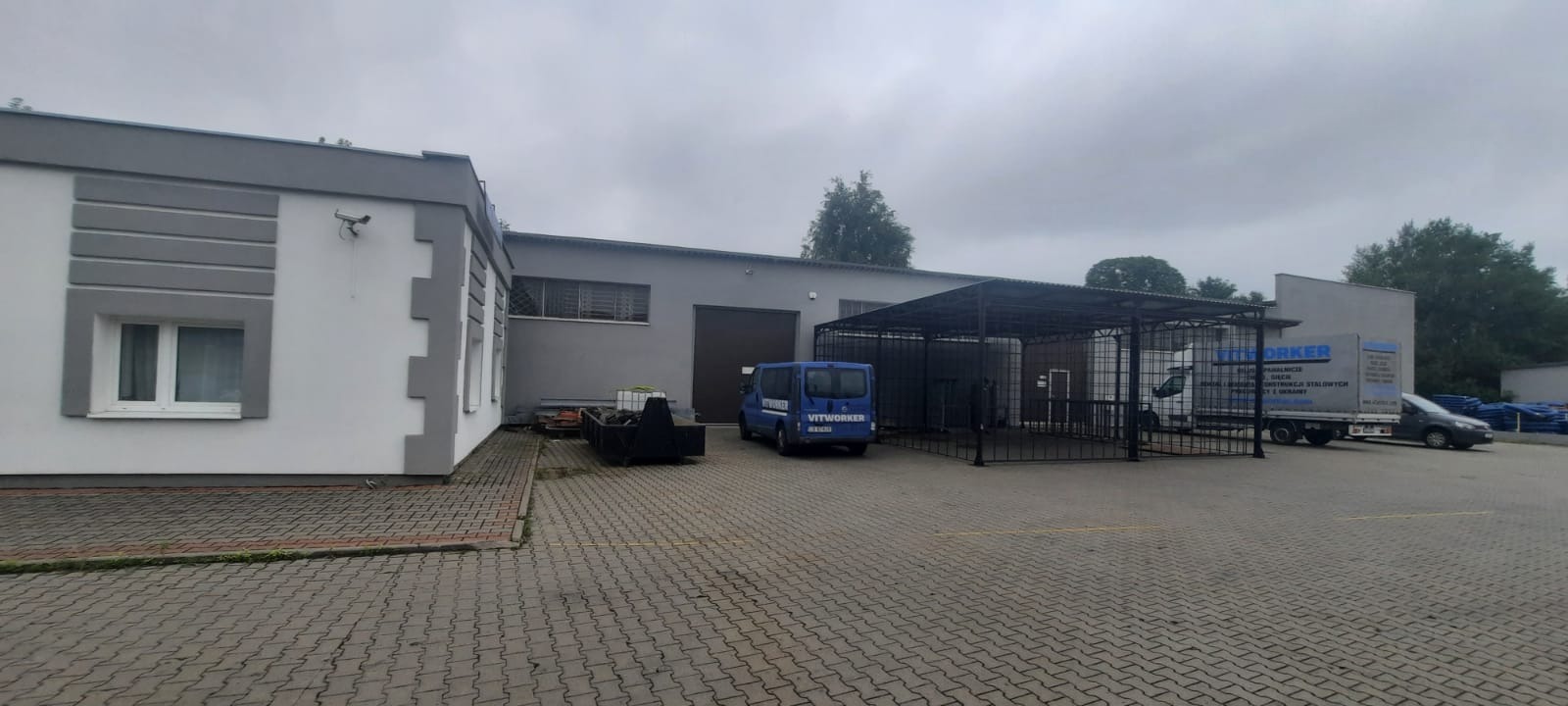 Budynek firmy z zaparkowanymi samochodami dostawczymi, w tle widoczna zadaszona konstrukcja.