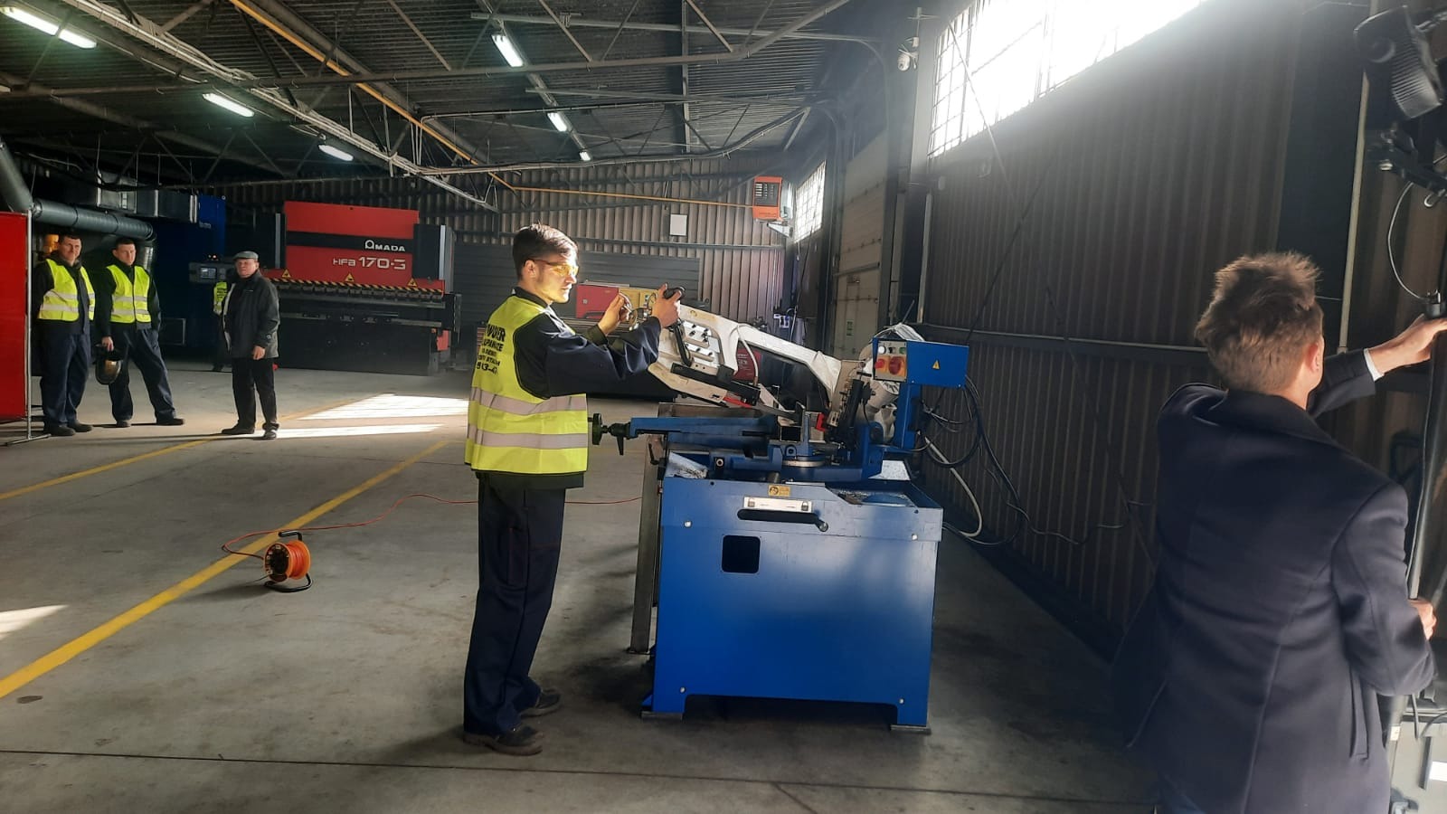 Pracownik w kamizelce odblaskowej obsługuje piłę taśmową do metalu w hali produkcyjnej, w tle inni pracownicy i maszyna do obróbki blachy.