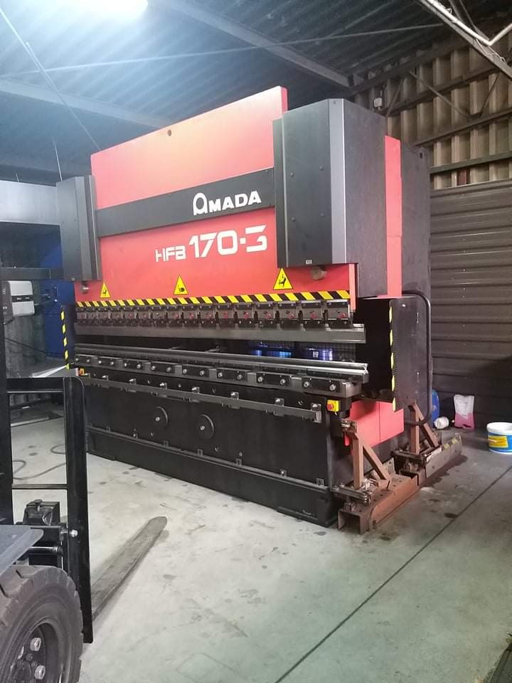 Czerwono-czarna prasa krawędziowa AMADA HFB 170-3 w hali produkcyjnej, widoczne ostrzeżenie o niebezpieczeństwie, obok wózek widłowy.