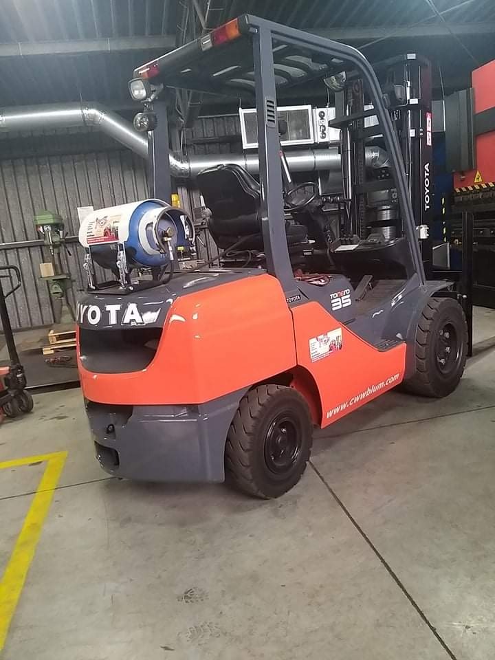 Szaro-pomarańczowy wózek widłowy Toyota z butlą gazową na tle hali produkcyjnej.