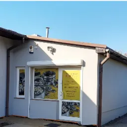 Fasada budynku sklepu z częściami samochodowymi Rombor z naklejonymi grafikami łożysk na oknach i drzwiach wejściowych, widoczny system alarmowy i oświetlenie zewnętrzne.