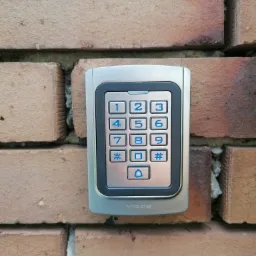 Szary panel numeryczny domofonu z niebieskimi cyframi, zamontowany na ceglanej ścianie zewnętrznej budynku.