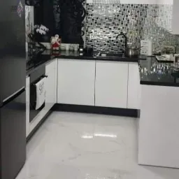 Nowoczesna kuchnia z białymi szafkami, czarnym blatem i mozaikową ścianą nad blatem. Czarna lodówka po lewej stronie i biała marmurowa podłoga.