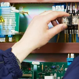 Elektryk w żółtym kasku instaluje przekaźnik w rozdzielnicy elektrycznej, widoczne bezpieczniki i okablowanie.