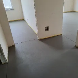 Szara, gładka posadzka jastrychowa w surowym stanie, zabezpieczona taśmą przy ścianach w nowym budynku z widocznymi instalacjami elektrycznymi.