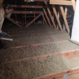 Pracownik w masce ochronnej rozpyla izolację z dyszy na drewnianą konstrukcję poddasza, widoczne przewody wentylacyjne pod sufitem.