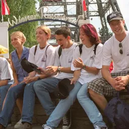 Grupa młodych ludzi siedzi razem na murku, ubrani w białe koszulki z logo, niektóre osoby w czapkach, w tle widać wiedeński Prater z charakterystycznym diabelskim młynem.