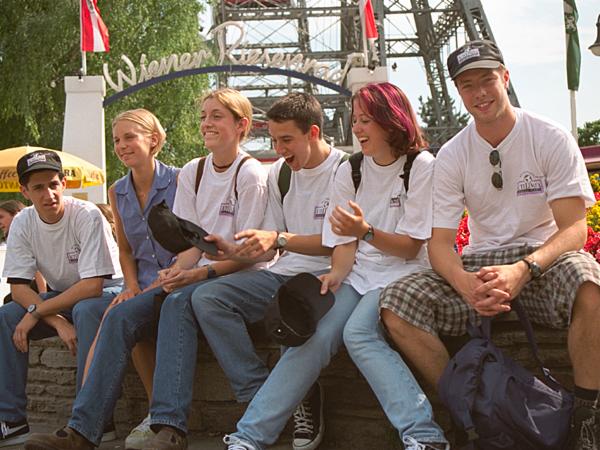 Grupa młodych ludzi siedzi razem na murku, ubrani w białe koszulki z logo, niektóre osoby w czapkach, w tle widać wiedeński Prater z charakterystycznym diabelskim młynem.
