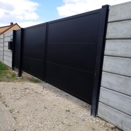 ABRAM zakład handlowo-usługowy - Nowoczesna, przesuwna brama wjazdowa w kolorze czarnym, z poziomymi przetłoczeniami, zamontowana między betonowymi przęsłami ogrodzenia.