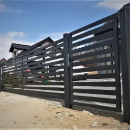 Nowoczesna, pozioma, metalowa brama wjazdowa przesuwna w kolorze grafitowym, widoczna z zewnątrz posesji, z domem w tle i fragmentami samochodów za bramą.