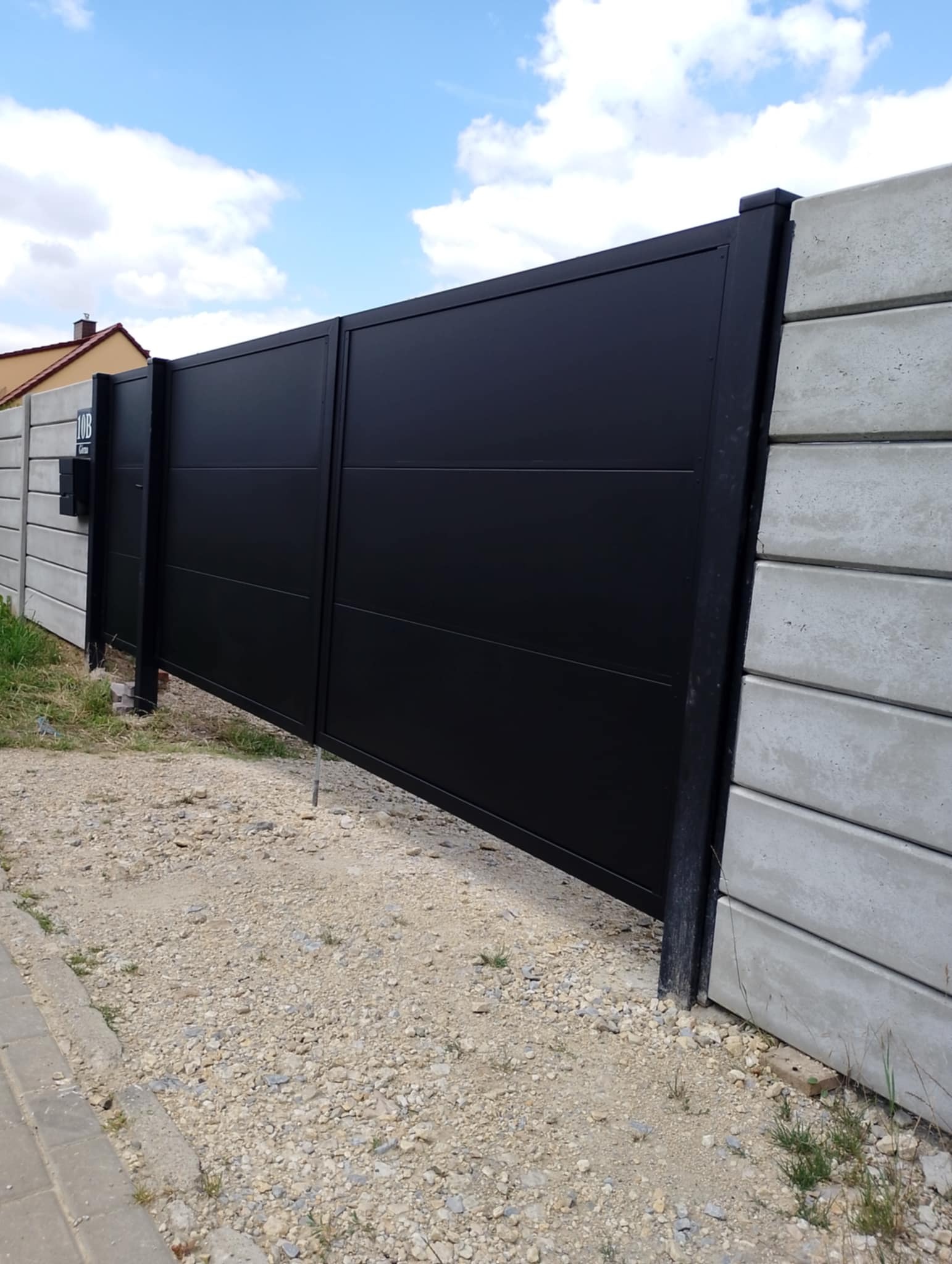 Nowoczesna, przesuwna brama wjazdowa w kolorze czarnym, z poziomymi przetłoczeniami, zamontowana między betonowymi przęsłami ogrodzenia.