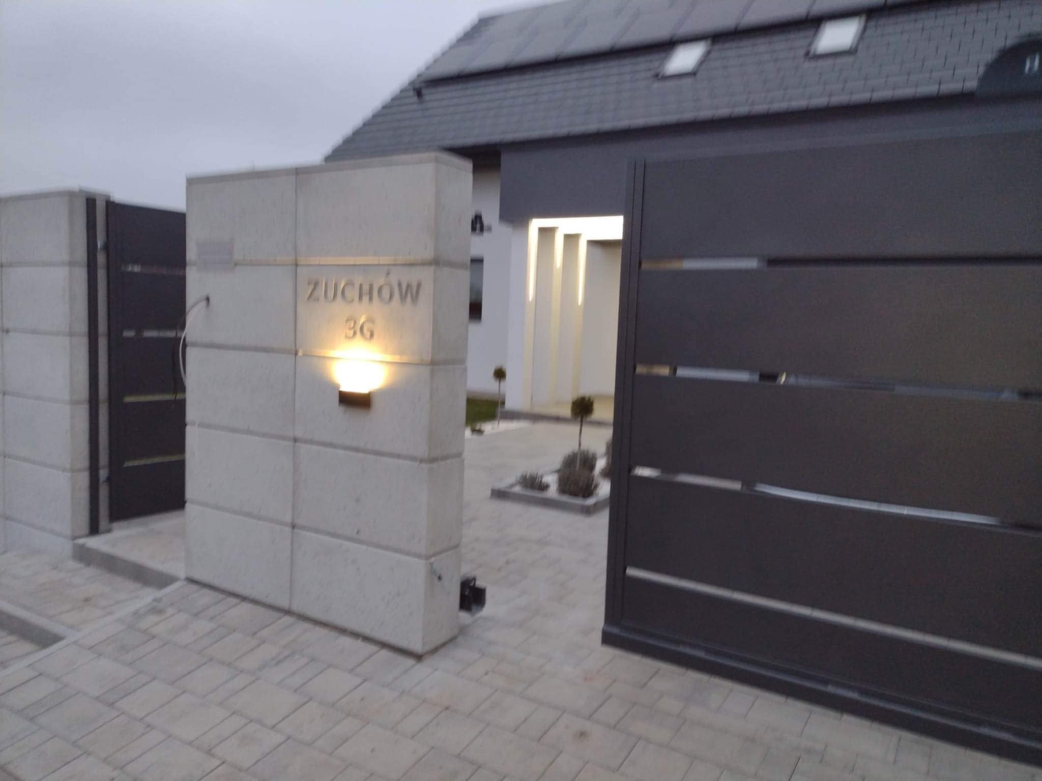 Nowoczesna, grafitowa brama wjazdowa z poziomymi panelami, zamontowana obok betonowego słupka z podświetlonym numerem domu 'Żuchów 36', na posesji z kostki brukowej.