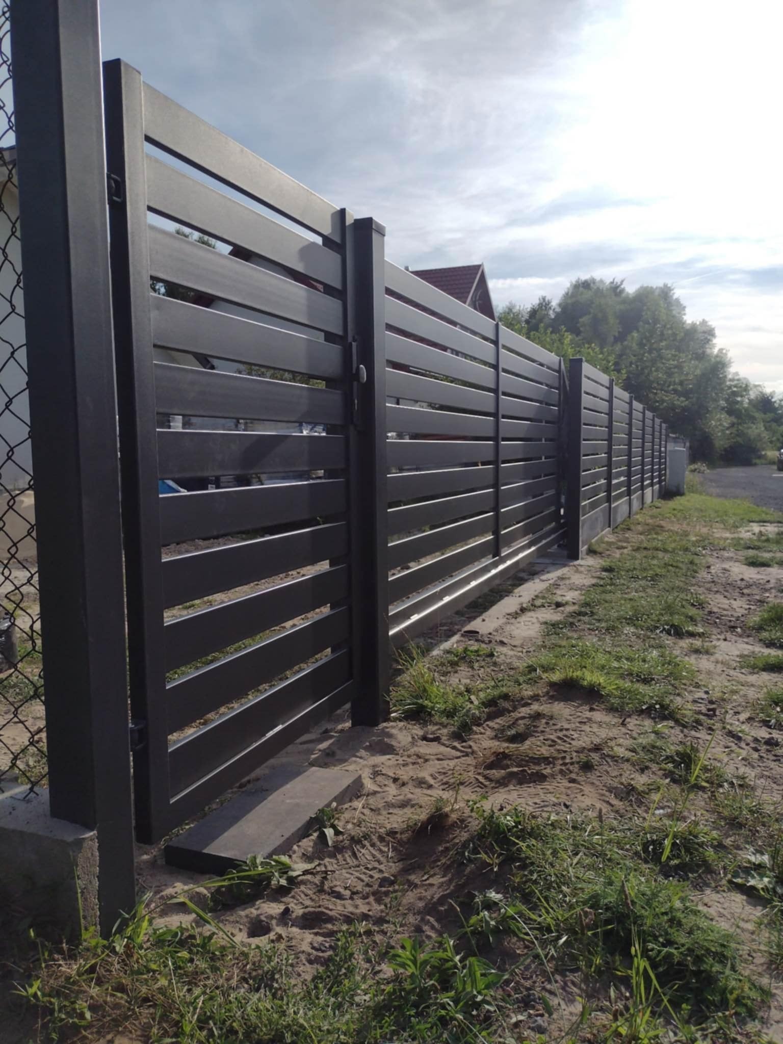 Nowoczesne, grafitowe ogrodzenie panelowe z poziomych profili, widoczne z boku, na tle trawy i drzew