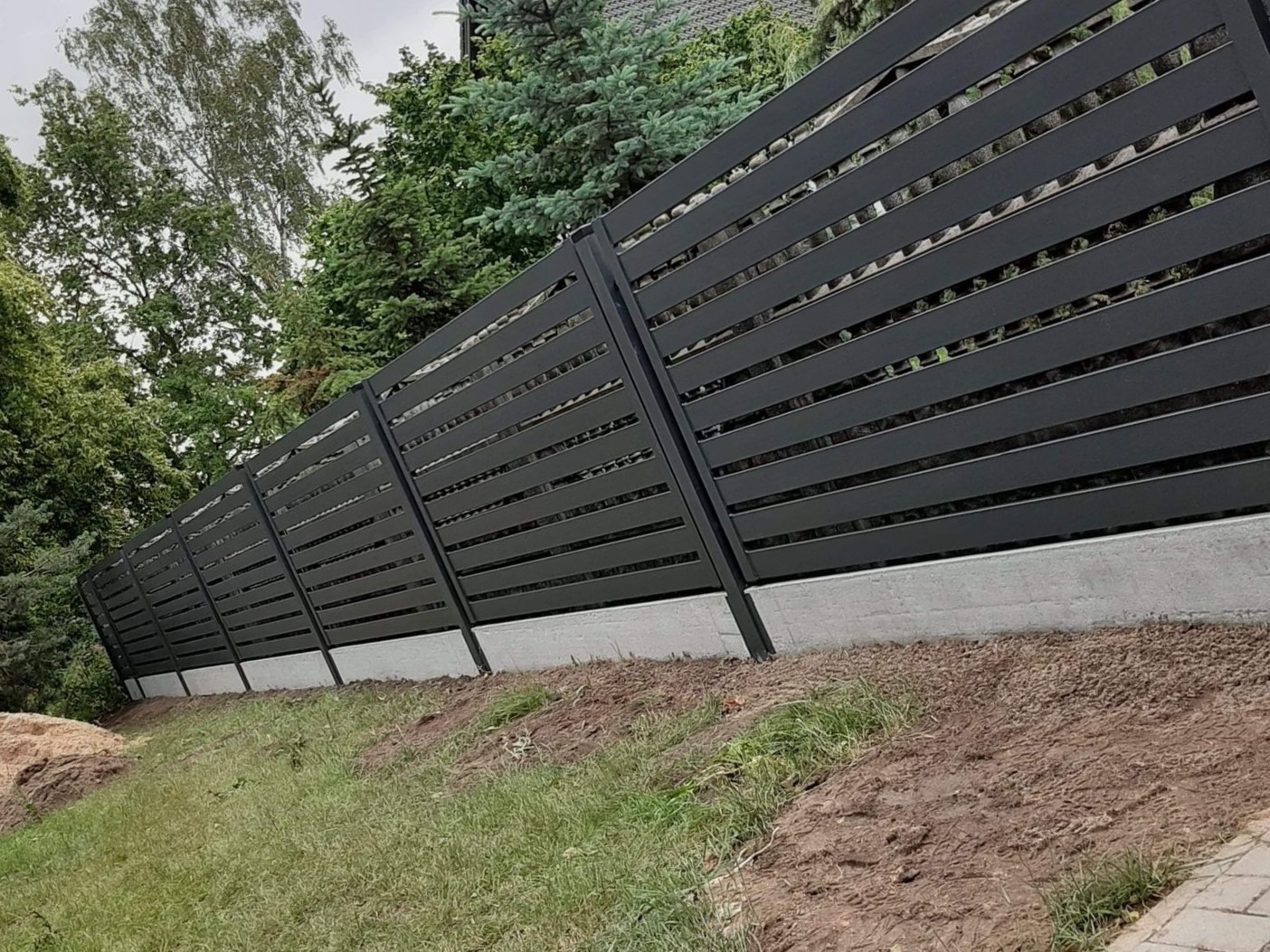 Długie, poziome, czarne przęsła tworzą nowoczesne ogrodzenie z metalowych profili, osadzone na betonowej podmurówce na pochyłym terenie.