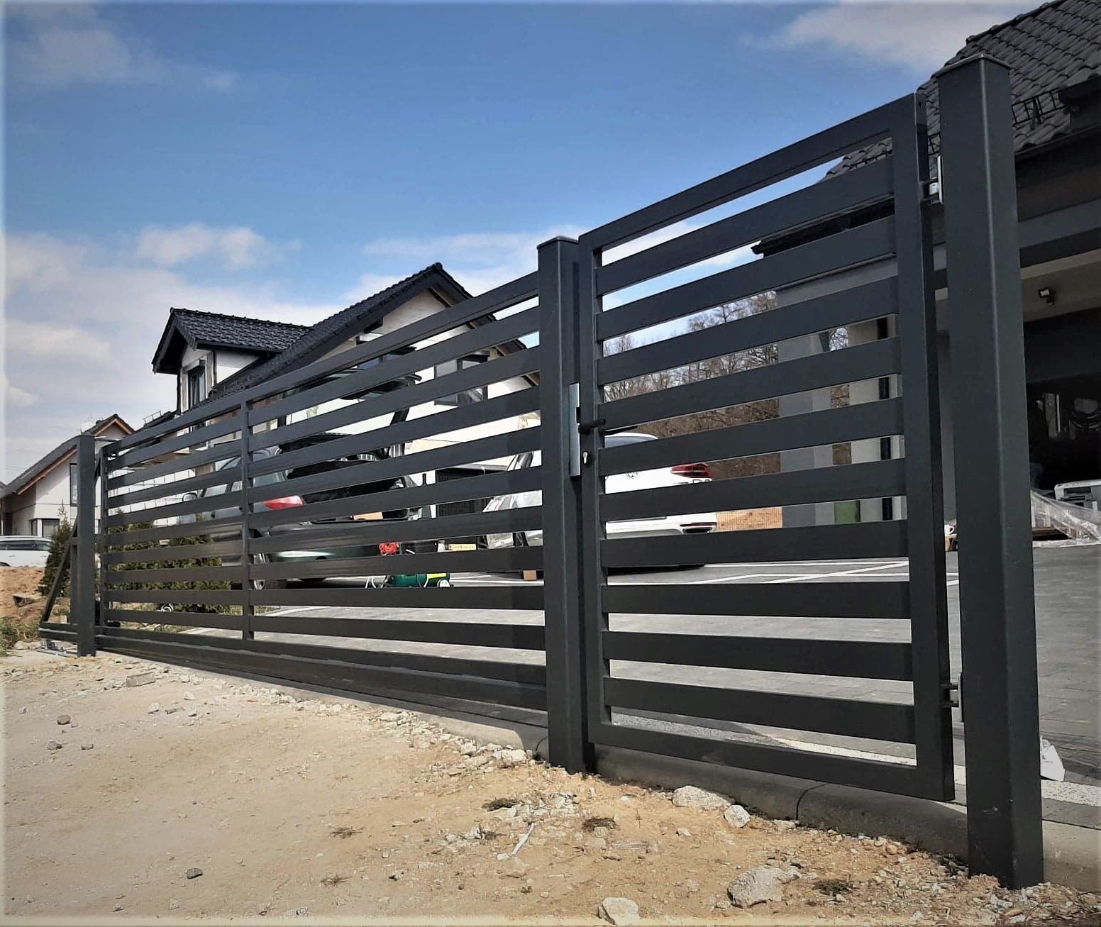 Nowoczesna, pozioma, metalowa brama wjazdowa przesuwna w kolorze grafitowym, widoczna z zewnątrz posesji, z domem w tle i fragmentami samochodów za bramą.