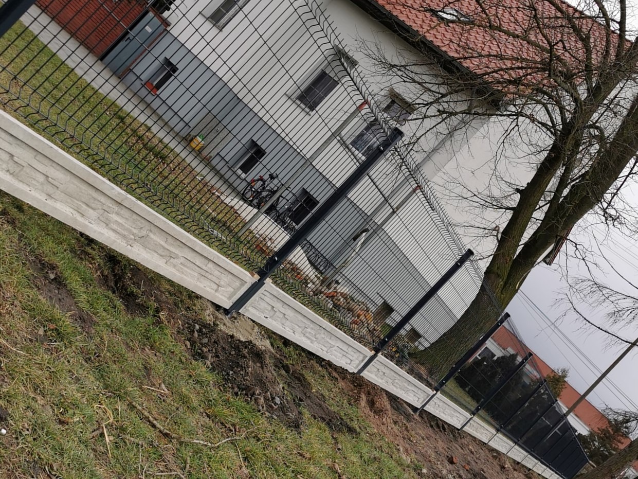 Nowoczesne ogrodzenie panelowe z podmurówką imitującą kamień, widoczne na tle budynku mieszkalnego i drzewa bez liści.