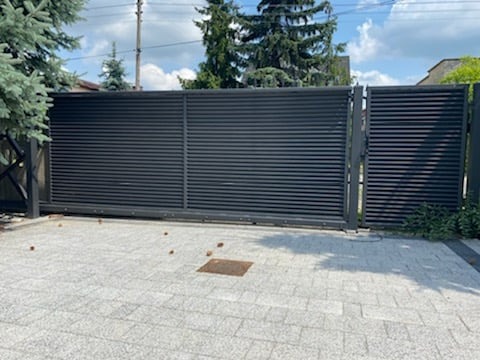 Nowoczesna, przesuwna brama wjazdowa z poziomych, czarnych profili, widoczna na tle zadbanego podwórza z kostki brukowej i iglaków.