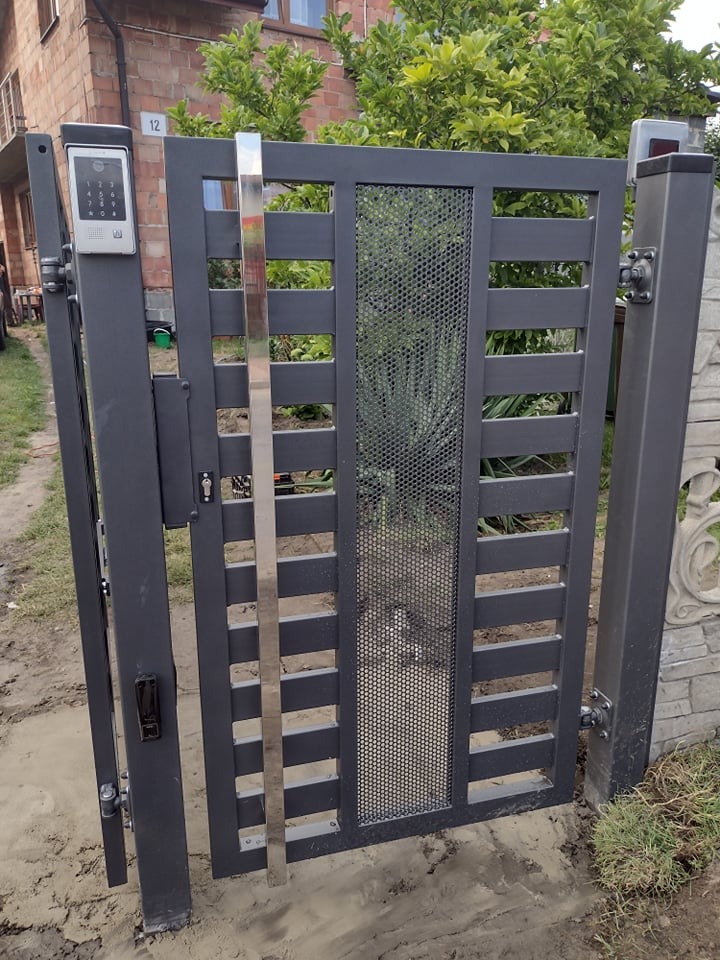 Nowoczesna, grafitowa brama wejściowa z perforowanym panelem centralnym i srebrnym, pionowym elementem dekoracyjnym, zamontowana przy posesji z domem z czerwonej cegły.