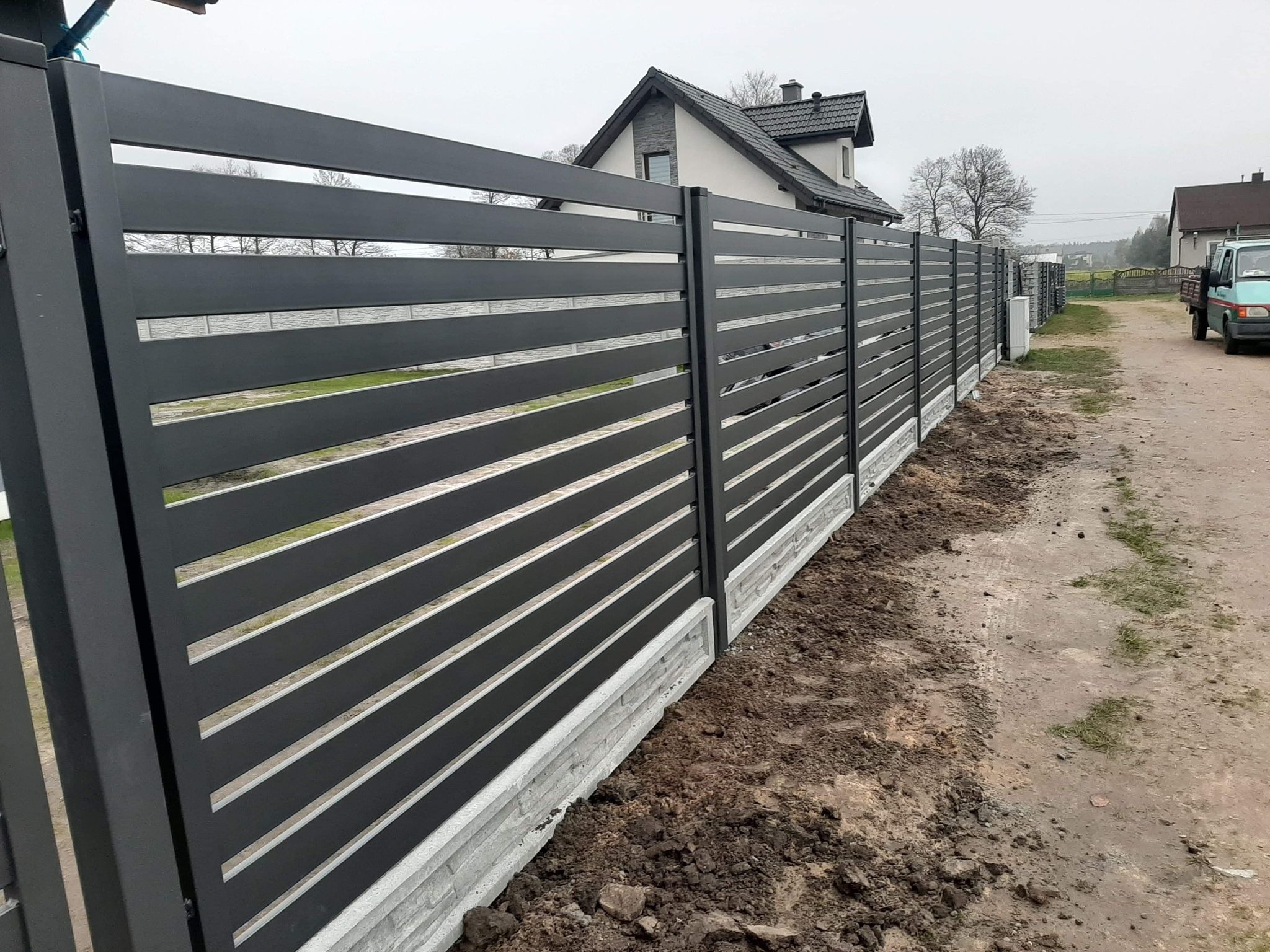 Nowoczesne, poziome, szare przęsła ogrodzeniowe na betonowej podmurówce, widok z zewnątrz posesji, w tle dom jednorodzinny i zaparkowany samochód dostawczy.