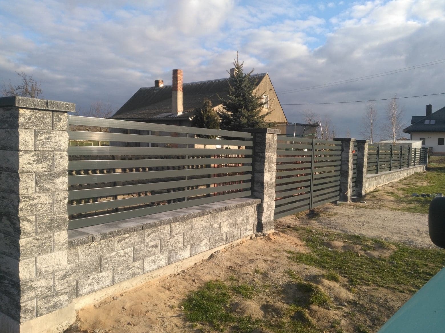 Nowoczesne ogrodzenie z poziomych, szarych metalowych profili, osadzone na murku i słupkach z szarej kostki łupanej, na tle domów i pochmurnego nieba.