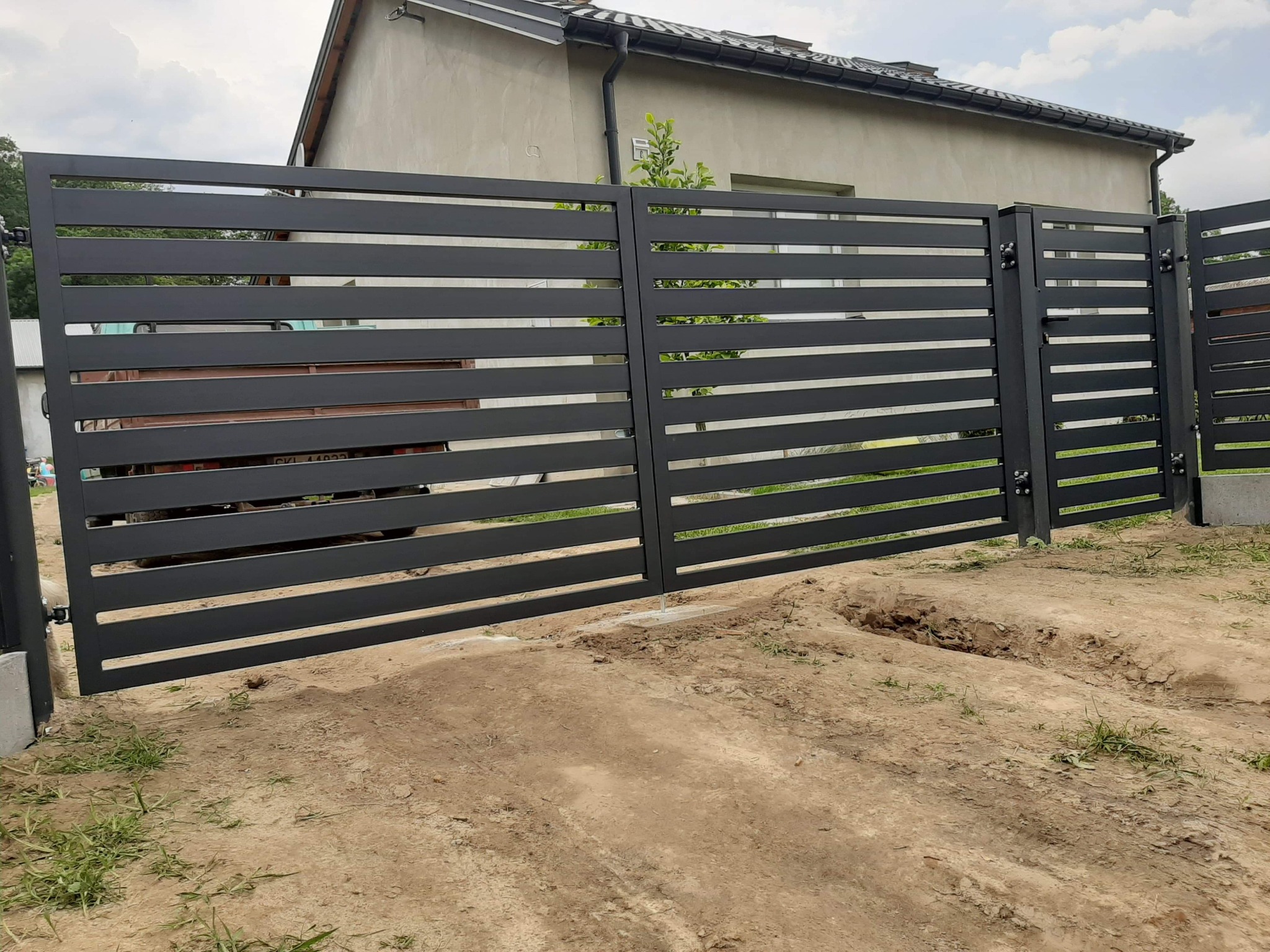 Nowoczesna, pozioma brama wjazdowa i ogrodzenie z metalowych profili w kolorze grafitowym, widoczny fragment posesji w tle, teren wokół bramy w trakcie prac ziemnych.