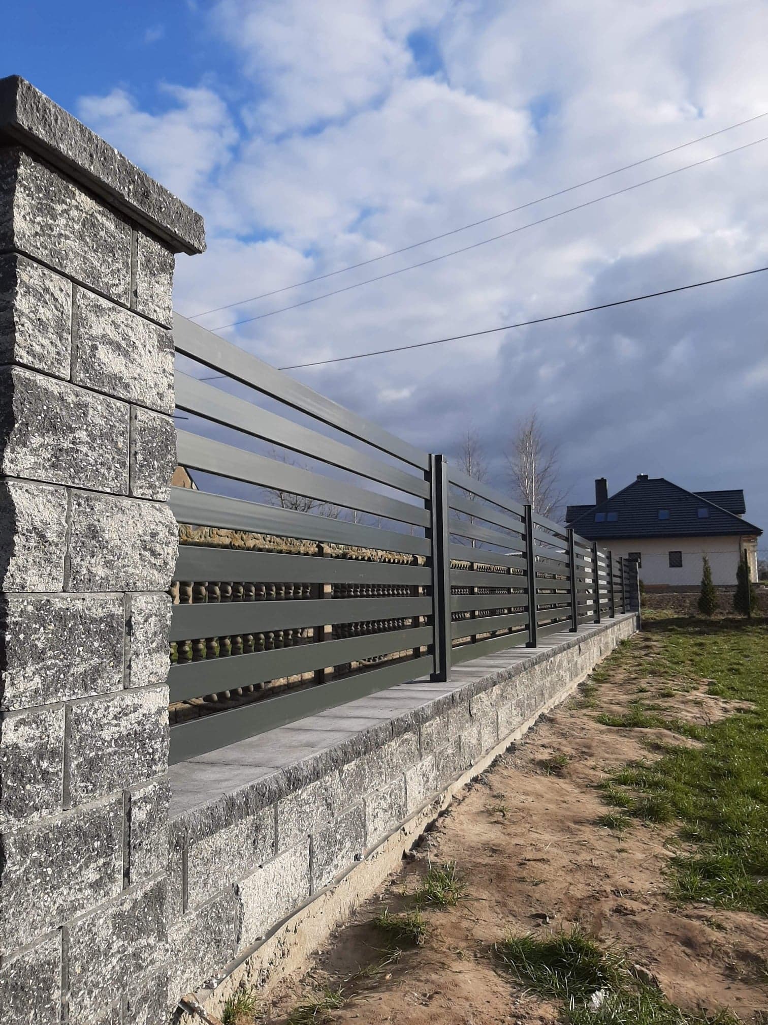 Nowoczesne ogrodzenie poziome z metalowymi przęsłami w kolorze grafitowym, osadzone na murku z szarej kostki łupanej, w tle dom jednorodzinny pod pochmurnym niebem.